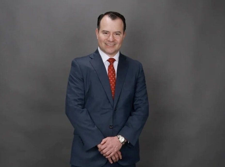 Man in a blue suit with a red tie standing against a gray background, hands together.